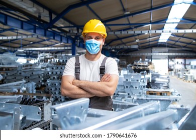Portrait of factory workman in uniform and hardhat wearing face mask in industrial production plant. People working during COVID-19 pandemic.