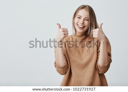 Portrait of fair-haired beautiful female student or customer with broad smile, looking at the camera with happy expression, showing thumbs-up with both hands, achieving study goals. Body language