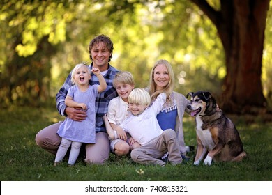 A Portrait Of A Family Of 5 Blonde Haired, Caucasian People, Sitting Outside Under The Trees With Their Adopted Pet German Shepherd Mix Breed Dog.