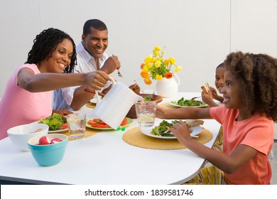 Portrait Of A Family Of Four At Dinner Table With Mother Pouring Water In Glass Of Daughter.