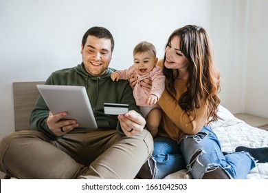 Portrait Of A Family On The Bed Looking At The Tablet With Their Baby Girl And Shopping Online With A Credit Card - Father, Mother And Little Daughter Have Fun Together - Intimacy Moment