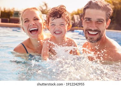 Portrait Of Family With Young Son Having Fun On Summer Vacation Splashing In Outdoor Swimming Pool