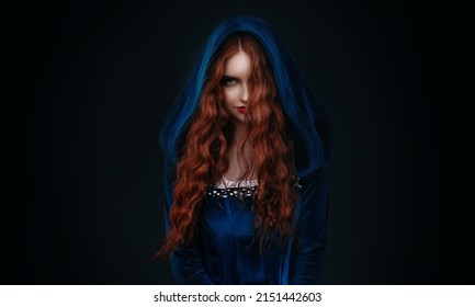 Portrait fantasy gothic red-haired woman witch. Vampire girl in blue medieval dress, vintage old historical style hood on head. Black background. Red lips, hair flying soar in wind. halloween costume