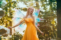 Portrait Fantasy Woman Blonde Forest Fairy. Elf Girl Fashion Model In Bright Yellow Dress, Butterfly Wings.  Walks In Summer Nature. Green Spring Tree, Wood, Sun Light Magic Radiance. Long Hair.