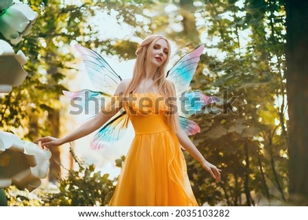 Portrait fantasy woman blonde forest fairy. Elf girl fashion model in bright yellow dress, butterfly wings.  walks in summer nature. Green spring tree, wood, sun light magic radiance. Long hair.