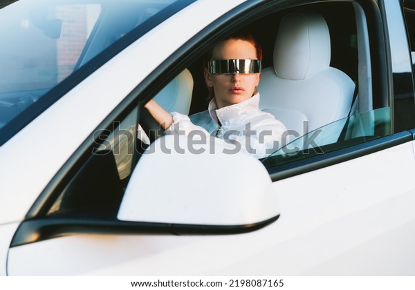 Portrait\
of fashion cyberpunk girl driving a white supercar. Red hair woman\
with futuristic eyeglasses sits by the car steering wheel. Future\
technology concept. Selective focus, copy\
space.