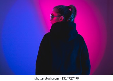 Portrait Of Fashion Young Girl In A Black Sweater With A Hood And Round Sunglasses In Red And Blue Neon Light In The Studio