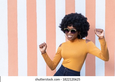 Portrait of fashionable African American happy woman with curly hairstyle, smiles broadly, exclaims with joy, wearing stylish sunglasses, enjoying dancing, posing on striped background outdoors.