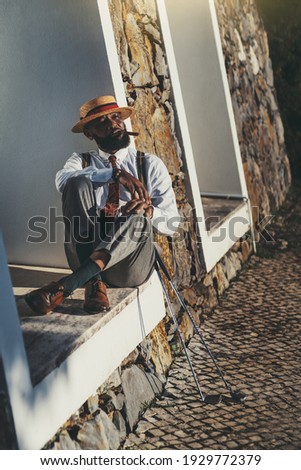 A portrait of a fashionable black guy in a hat and an elegant outfit sitting on a sill outdoors and going to smoke a cigar after a good golf play, two golf clubs of different type near him
