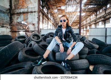 Portrait Of Fashionable Blonde Girl Wearing A Rock Black Style Outdoors,  Sits On Old Car Tires In An Abandoned Factory