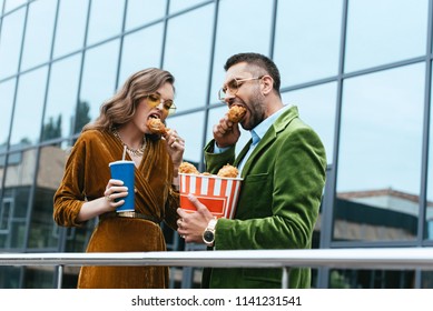 Portrait Of Fashionable Couple In Velvet Clothing Eating Fried Chicken Legs On Street