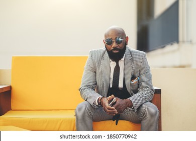 A portrait of a fashionable handsome mature bald black guy with a beard and in an elegant plaid costume with a necktie, in round sunglasses, sitting on an outdoor soft bench of a vivid yellow color