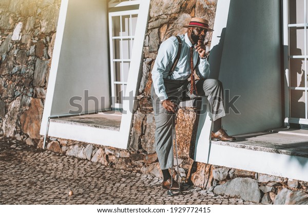 A portrait of a fashionable mature black guy with\
a beard and in a hat and an elegant costume waiting for his friends\
before a golf play while standing with a cigar in hand next to the\
building facade