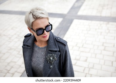 Portrait of fashionable mature woman with short blonde hair wearing stylish gray wool coat with lapels and shoulder straps, accessories brooch and sunglasses - Powered by Shutterstock