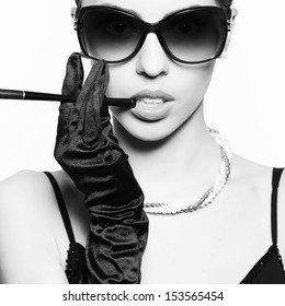 Portrait of a fashionable model in vintage sunglasses with a cigarette holder. Perfect skin. Great pearl accessories. Retro style. Close up. Black & white (monochrome) studio shot