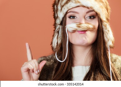 Portrait Of Fashionable Pretty Woman In Fur Winter Cap Hat With Funny Mustache And Finger Showing Copy Space On Orange Background In Studio.