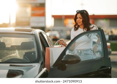 Portrait of fashionable smiling young woman with paper shopping bags in hands entering her car while standing on a parking area on city street downtown. Happy shopper girl entering car after shopping. - Powered by Shutterstock