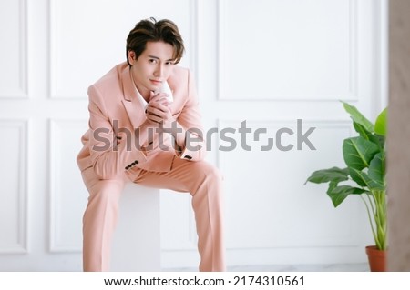Portrait of fashionable stylish business young Asian man in light pink suit sitting on white cubes and looking at camera at office with scandinavian decor. Copy space. Korean cool man freelance work.