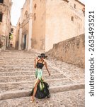 Portrait of fashionable woman walking sideways in beautiful old stone city town Calvi in island Corsica, France. Model wears elegant  feminine outfit, hat and silk skirt showing her long leg, while wa