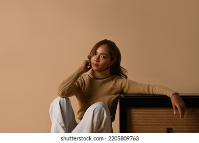 Portrait Of Fashionable Woman Wearing Trendy Sweater Sitting Near Vintage Radio, Studio Shot On Beige Background