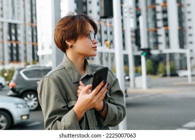 Portrait Of A Fashionable Young Asian Woman In Stylish Glasses And With A Short Haircut Holding A Smartphone And Looking Away While Standing On Street In City.