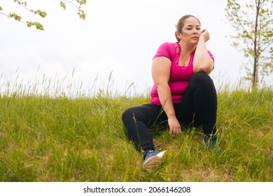 Portrait of fat attractive young woman sitting on green grass in city park looking at camera. Confident overweight female sits in nature having leisure activity outdoors in alone.
