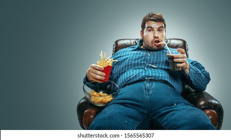 Portrait of fat caucasian man wearing jeanse and whirt sitting in a brown armchair on gradient grey background. Watching TV drinks cola, eats chips, fried potato, laughting. Overweight, carefree.