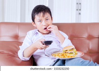 Portrait Of A Fat Little Boy Eating Junk Foods While Sitting On The Couch And Watching TV. Shot At Home