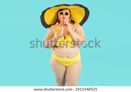 Portrait of fat woman in yellow swimsuit, beach hat and sunglasses drinking orange juice cocktail and looking cheerful at camera on blue background. Summer holiday trip and vacation concept.