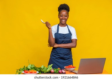 Portrait Of A Female African Chef Smiling. Young Black Chef Standing In Front Of A Table With Vegetables And Tomatoes On It, Also A Laptop, Holding A Spoon, Smiling