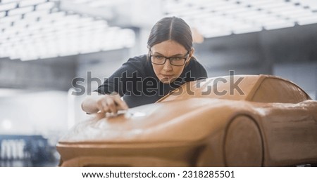 Portrait of a Female Automotive Designer Sculpting a 3D Clay Model of a New Production Car. Young Woman Using a Spatula to Carefully Trim the Surface of a Prototype Concept Vehicle