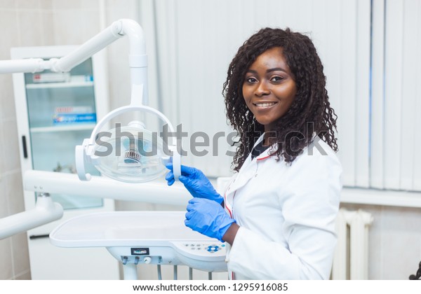 Portrait of Female black dentist in dental\
office. She standing at her office and she has beautiful smile.\
Modern medical\
equipment