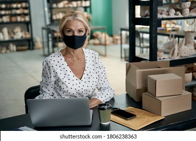 Portrait of female business owner or businesswoman wearing protective face mask using laptop and looking at camera while working in her art studio or craft pottery shop - Powered by Shutterstock