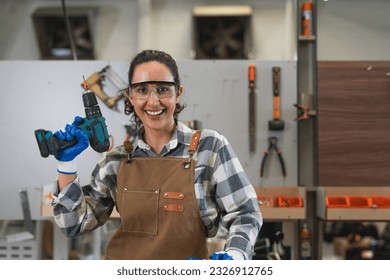 Portrait of female carpenter in apron smile and holding electric drill standing in carpentry workshop. Woman wearing glasses safety working with wood at furniture factory - Powered by Shutterstock