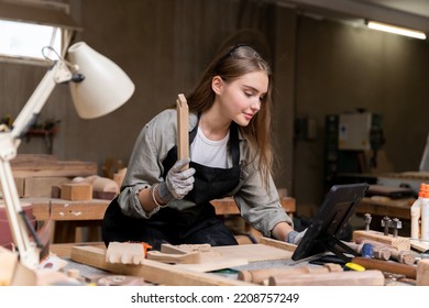 Portrait Of A Female Carpenter Looking At Designs On A Laptop For Making Her Furniture In A Furniture Factory. With Many Tools And Wood