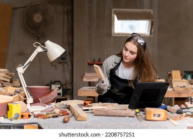 Portrait Of A Female Carpenter Looking At Designs On A Laptop For Making Her Furniture In A Furniture Factory. With Many Tools And Wood