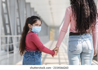 Portrait Of Female Child In Protective Medical Mask Walking With Mom At Airport, Little Girl Holding Mother's Hand And Turning At Camera, Traveling With Parent During Coronavirus Pandemic