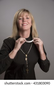 Portrait Of A Female Clergy Wearing A Black Shirt And Holding A Clerical Collar Which She Is Attaching To Her Shirt