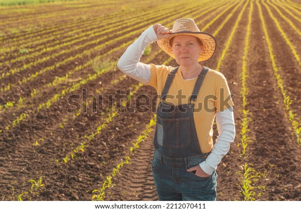 Portrait of female corn farmer in cultivated\
maize field wearing straw hat and jeans bib overalls and standing\
among young crop\
seedlings