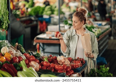 Portrait of female customer standing near market stall with paper bag in hands and shopping fresh organic tomato at farmer's market. Young shopper woman purchasing fresh products at food market. - Powered by Shutterstock