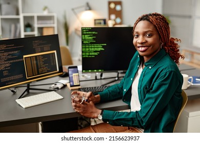 Portrait of female IT developer looking at camera and smiling against programming code on computer screen in office interior, copy space
