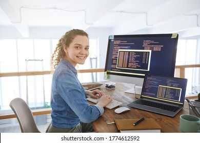 Portrait of female IT developer smiling at camera while typing on keyboard with black and orange programming code on computer screen and laptop in contemporary office interior, copy space