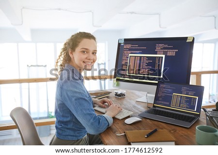 Portrait of female IT developer smiling at camera while typing on keyboard with black and orange programming code on computer screen and laptop in contemporary office interior, copy space