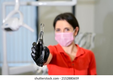 Portrait Of Female Doctor, Attractive Caucasian Armenian Ethnic Dentist Standing In Dental Office At Clinic, Smiling, Looking At Camera With Tooth Extraction Forceps, Tool For Removal Teeth Or Tooth