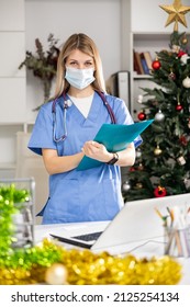 Portrait Of Female Doctor In Face Mask Working With Paperwork And Laptop In Office At Christmas