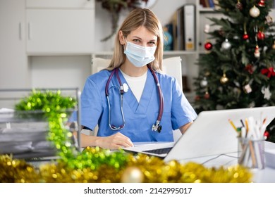 Portrait Of Female Doctor In Face Mask Working With Paperwork And Laptop In Office At Christmas