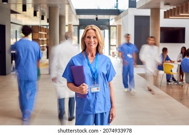 Portrait Of Female Doctor Wearing Scrubs Holding Patient Notes In Busy Hospital