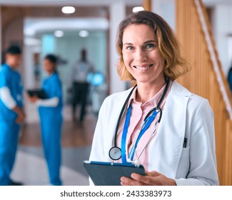 Portrait Of Female Doctor In White Coat And Digital Tablet In Hospital With Colleagues In Background - Powered by Shutterstock