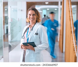 Portrait Of Female Doctor In White Coat And Digital Tablet In Hospital With Colleagues In Background - Powered by Shutterstock