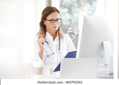 Portrait Of Female Doctor Writing On Clipboard While Sitting In Front Of Computer And Laptop At Doctors Office. 